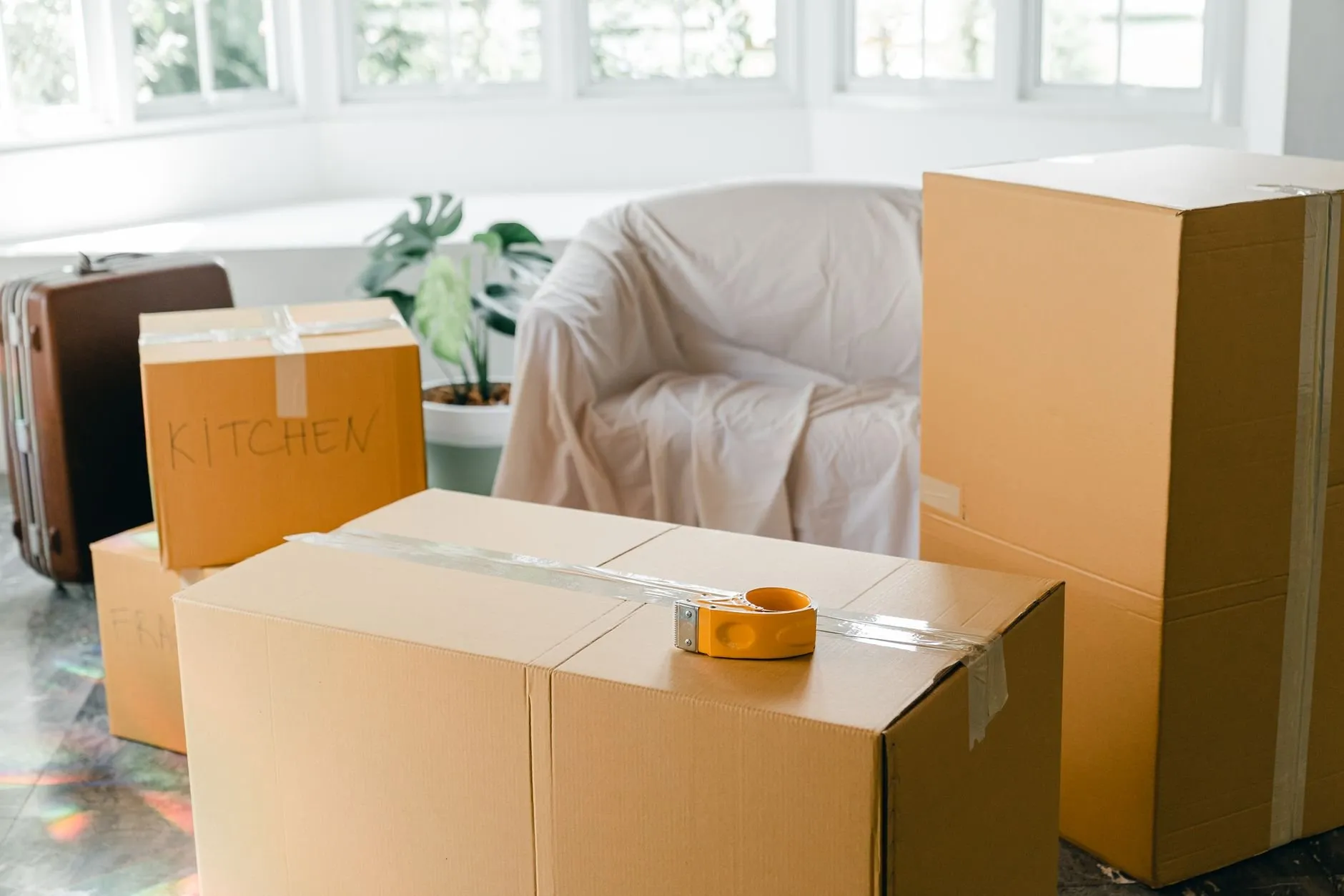 An armchair under a dust sheet surrounded by taped cardboard boxes and a suitcase