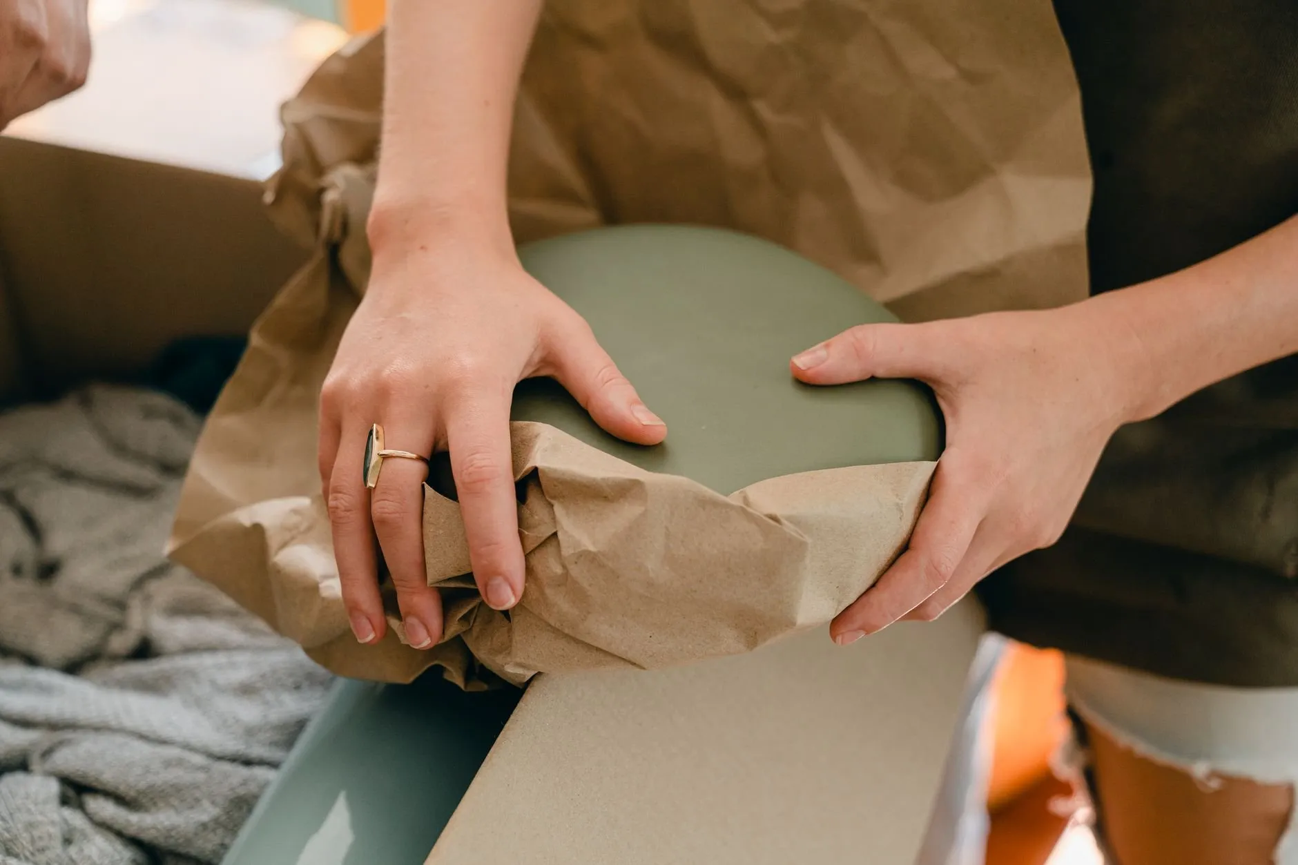 Hands wrapping a sage green vase in brown packing paper